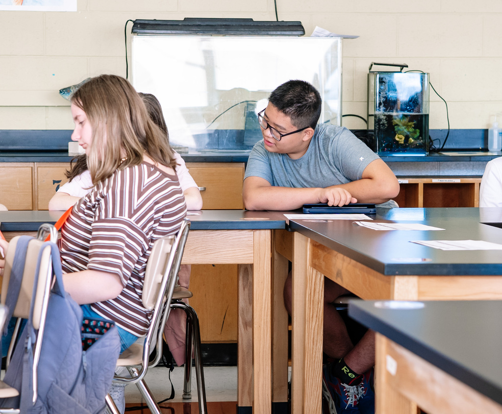 A. student looking at his friends schedule for the day while they are sitting at a table in a classroom. 