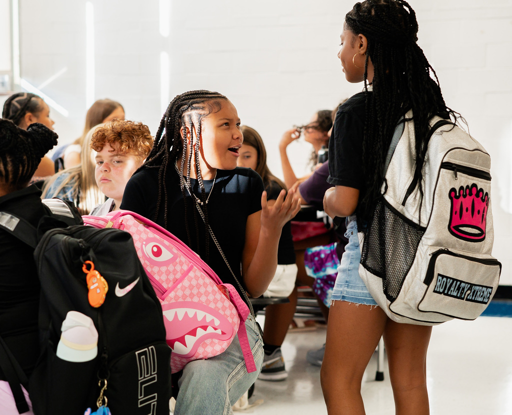 One student is sitting at a lunch table and is talking to another student who is tanding beside and is wearing a silver backpack that says Royalty Xtreme.