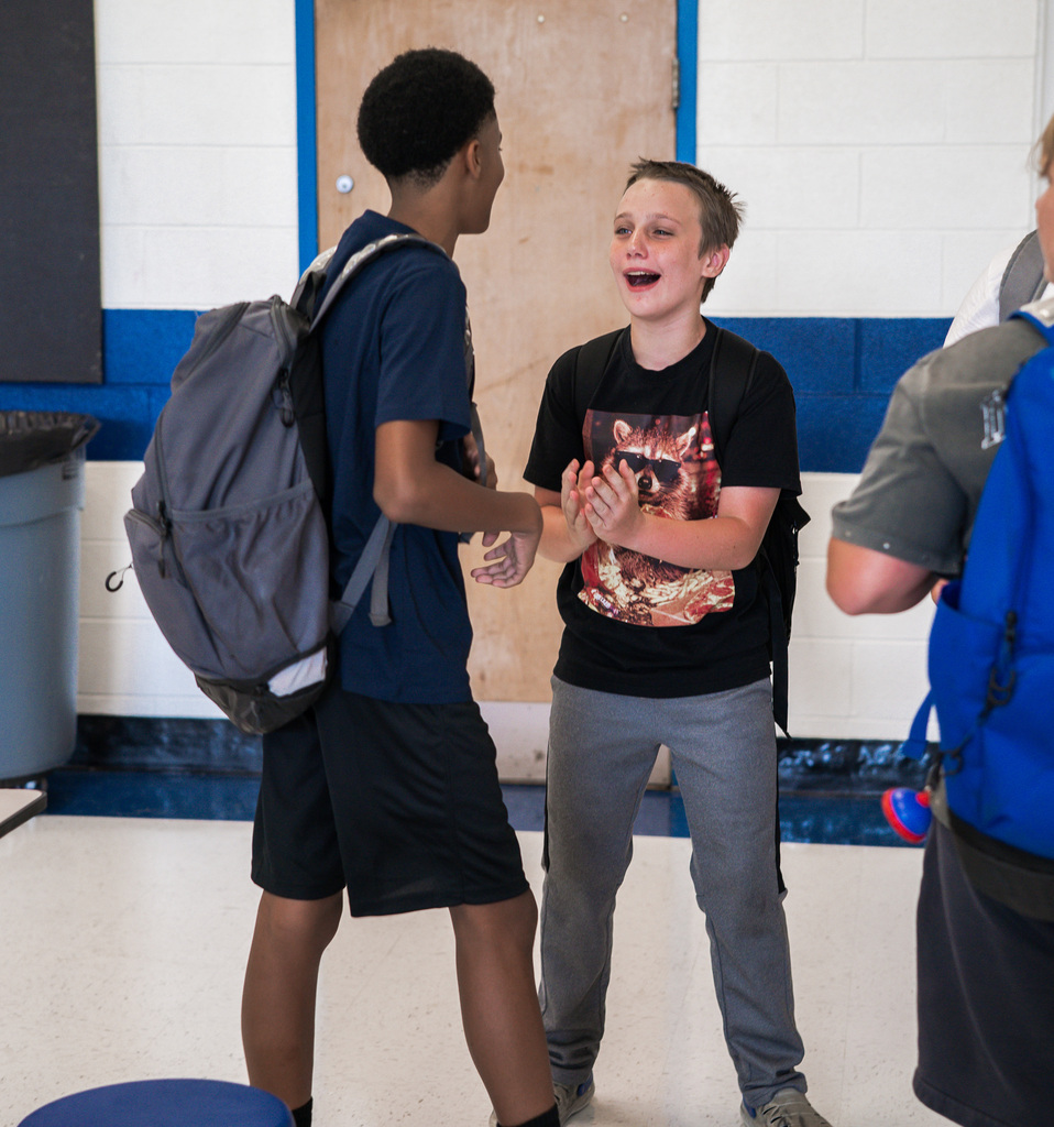 Two students are greeting each other while standing in the school cafeteria.