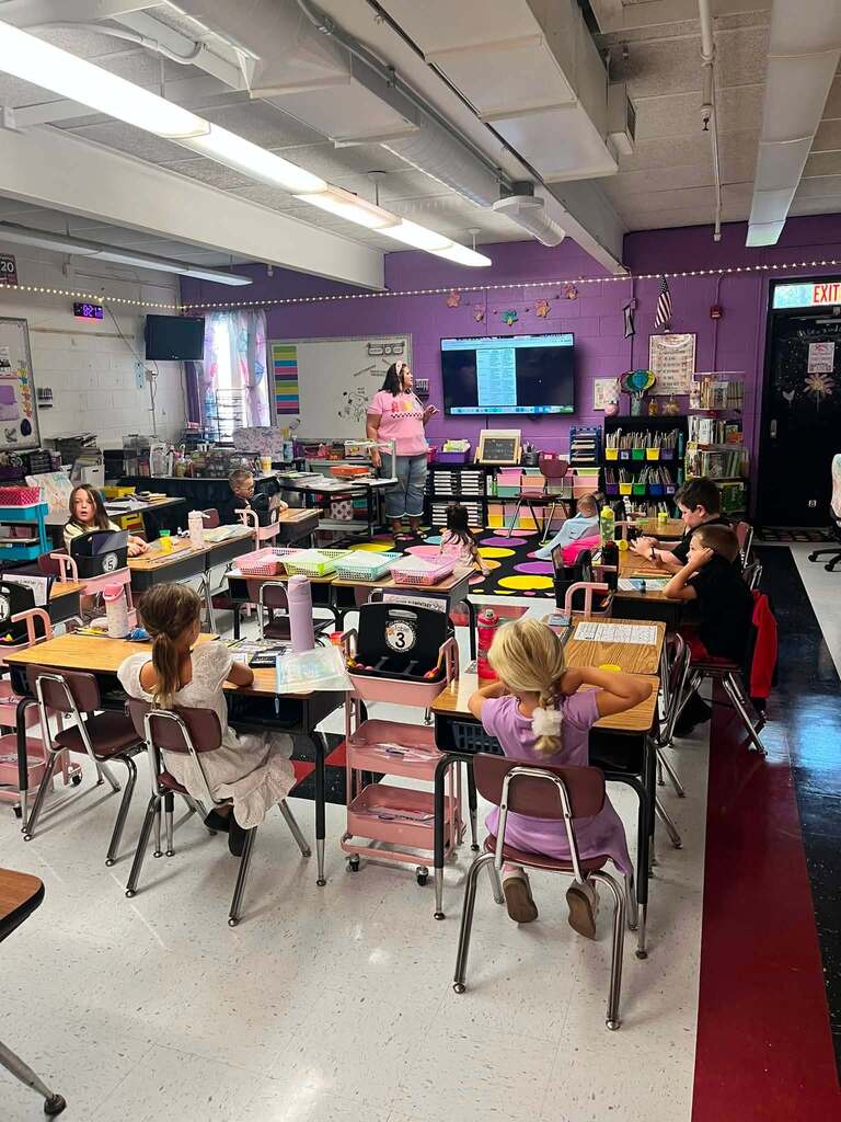 Students sitting at their desks in their classroom. The teacher is at the front of the room referencing something that is displayed on the TV.
