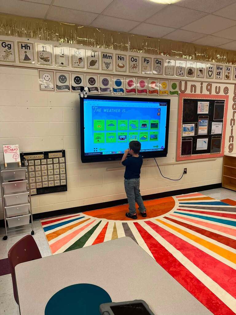 A young student standing at a TV in his classroom. The TV is displaying options for the weather outside and the student is looking to find the correct weather option to select.