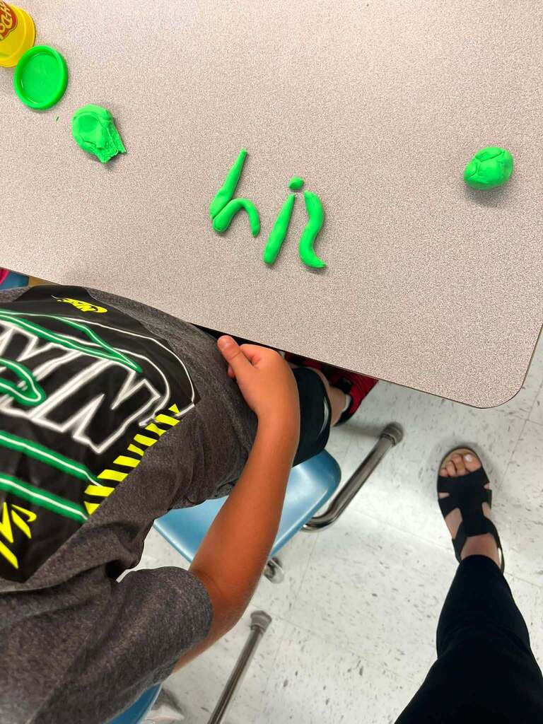The word Hi spelled out in green play-doh...the play-doh is on top of a student's desk.