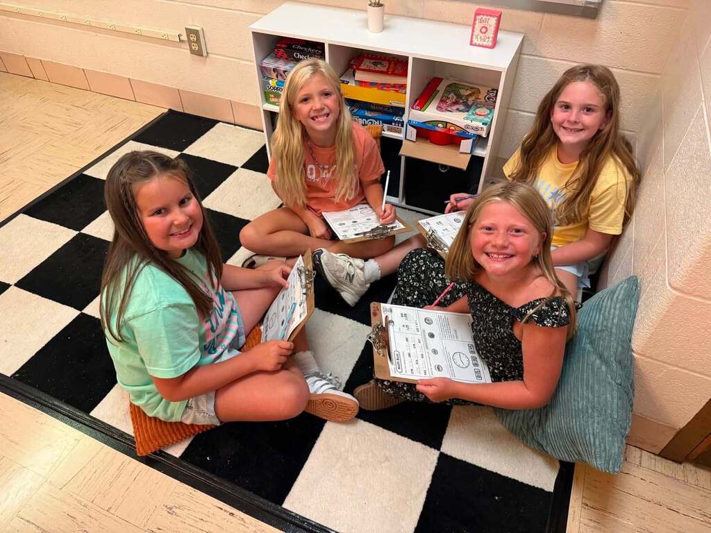 A group of four students are sitting on a black and white checkered mat in their classroom while doing a worksheet. The worksheet is being held by a clipboard. The girls are smiling at the camera.