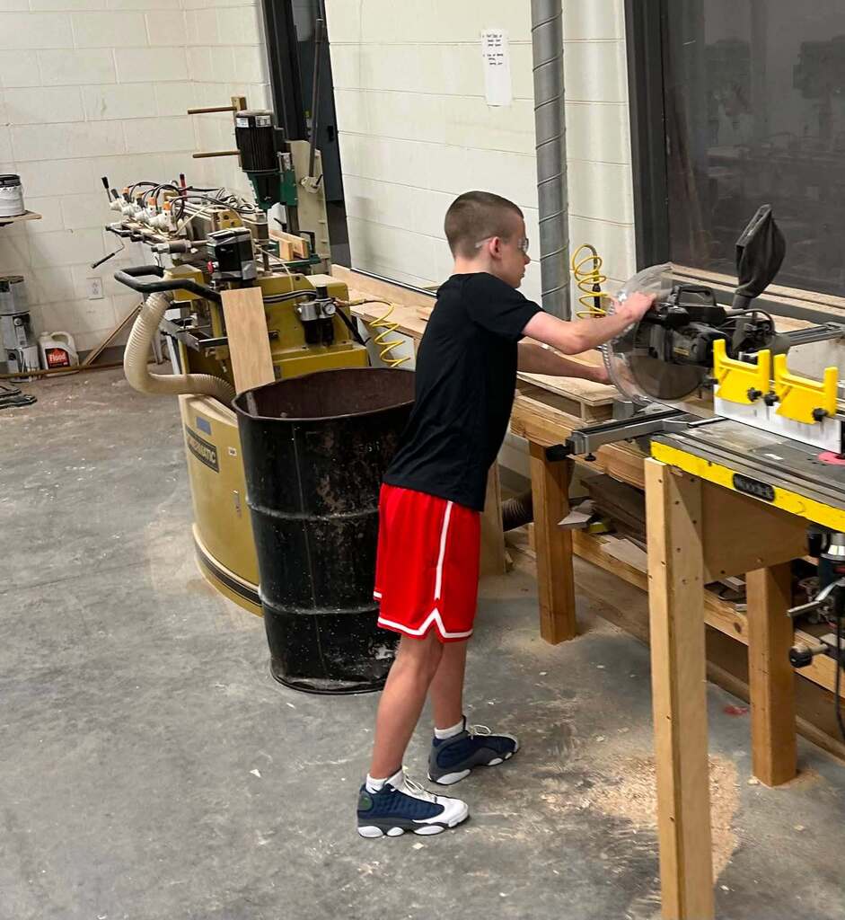 A high school student is using a compound miter saw to cut a piece of wood in shop class.