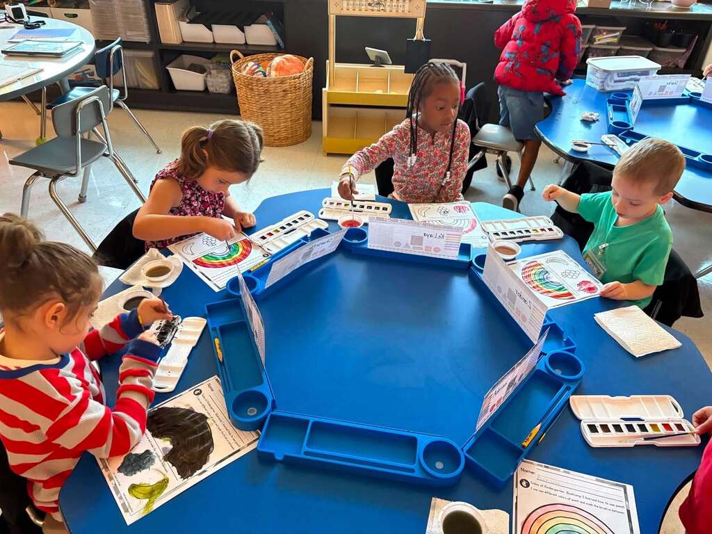 Young students sitting around a blue classroom table with painting supplies. They are painting a picture of a rainbow.