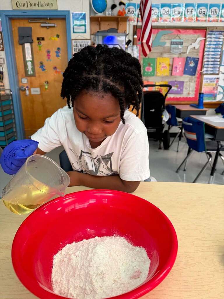 A young student, wearing blue latex gloves, is pouring vegetable oil into a red mixing bowl that has flower in it.