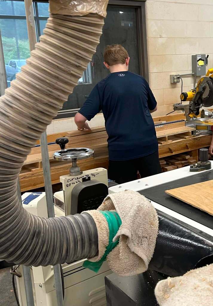 A high school student is measuring a piece of wood he is getting ready to cut. Behind the student is a CNC machine.