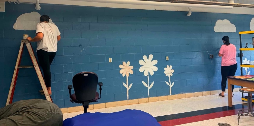 A student standing on a ladder and painting a cloud on a blue wall.