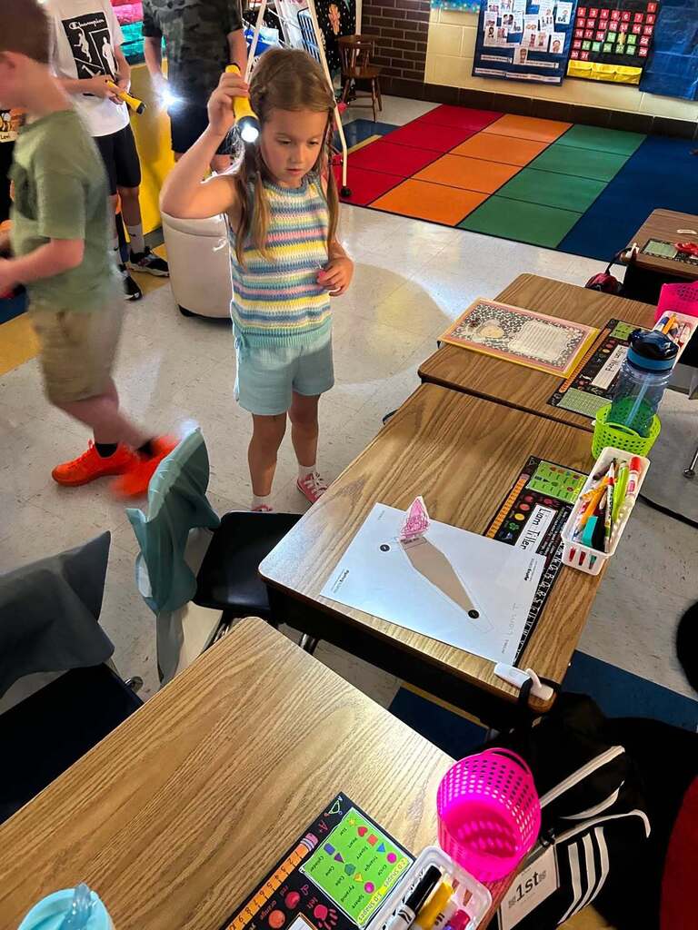A young student holding a flashlight by her head and shining it down to a paper cutout on her desk while doing a lesson about lights and shadows.