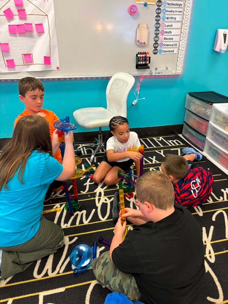 Students sitting on a an ABC rug in their classroom making towers together out of plastic tubes that connect together and are in different shapes and sizes.