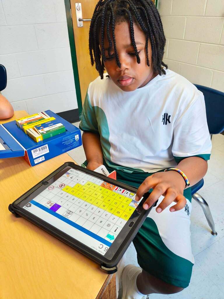A student sitting at a classroom table using his AAC device on his iPad during speech therapy.