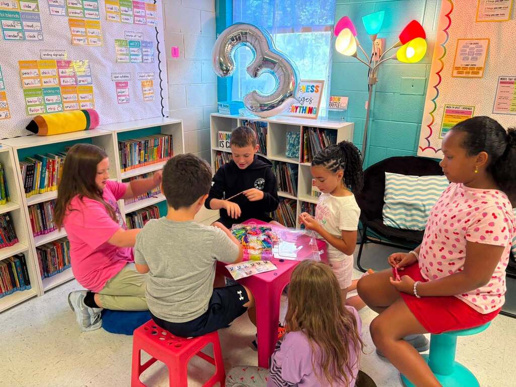 Students sitting around a small pink table in their classroom making bracelets out of different color beads and string.
