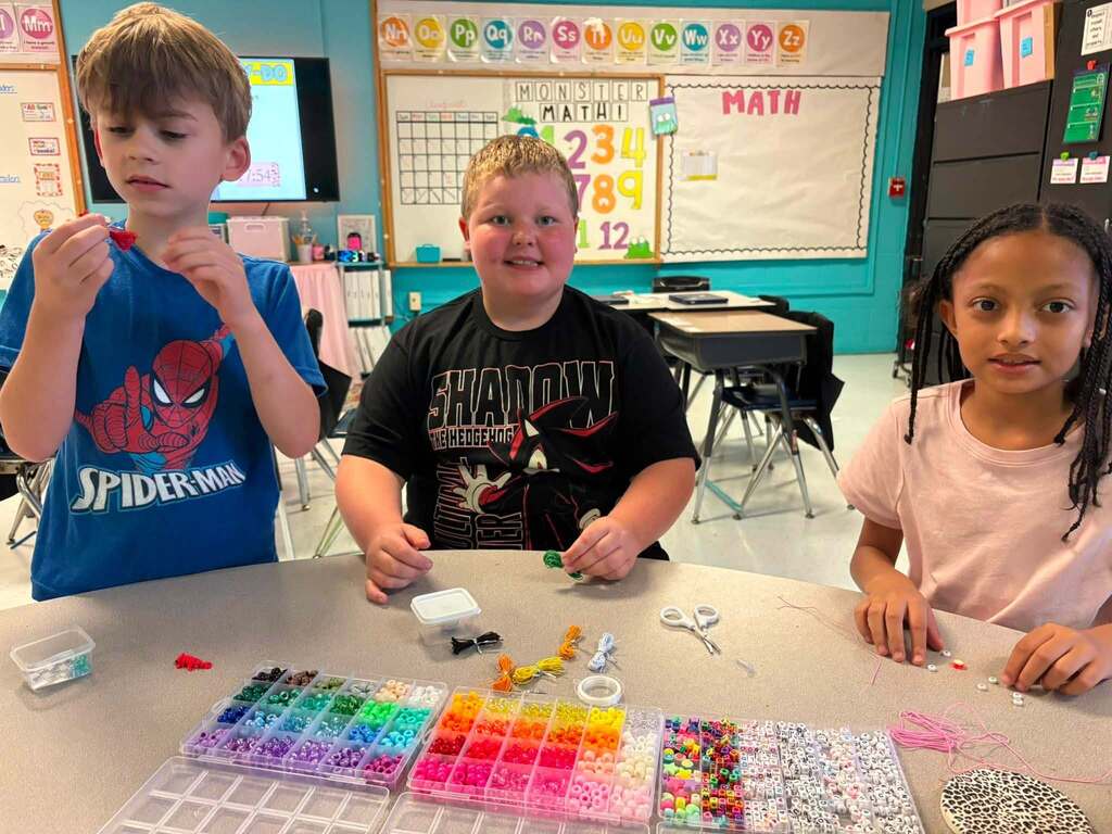 three students sitting at a classroom table and smiling at the camera. There are different color beads and string to make bracelets sitting on the table.