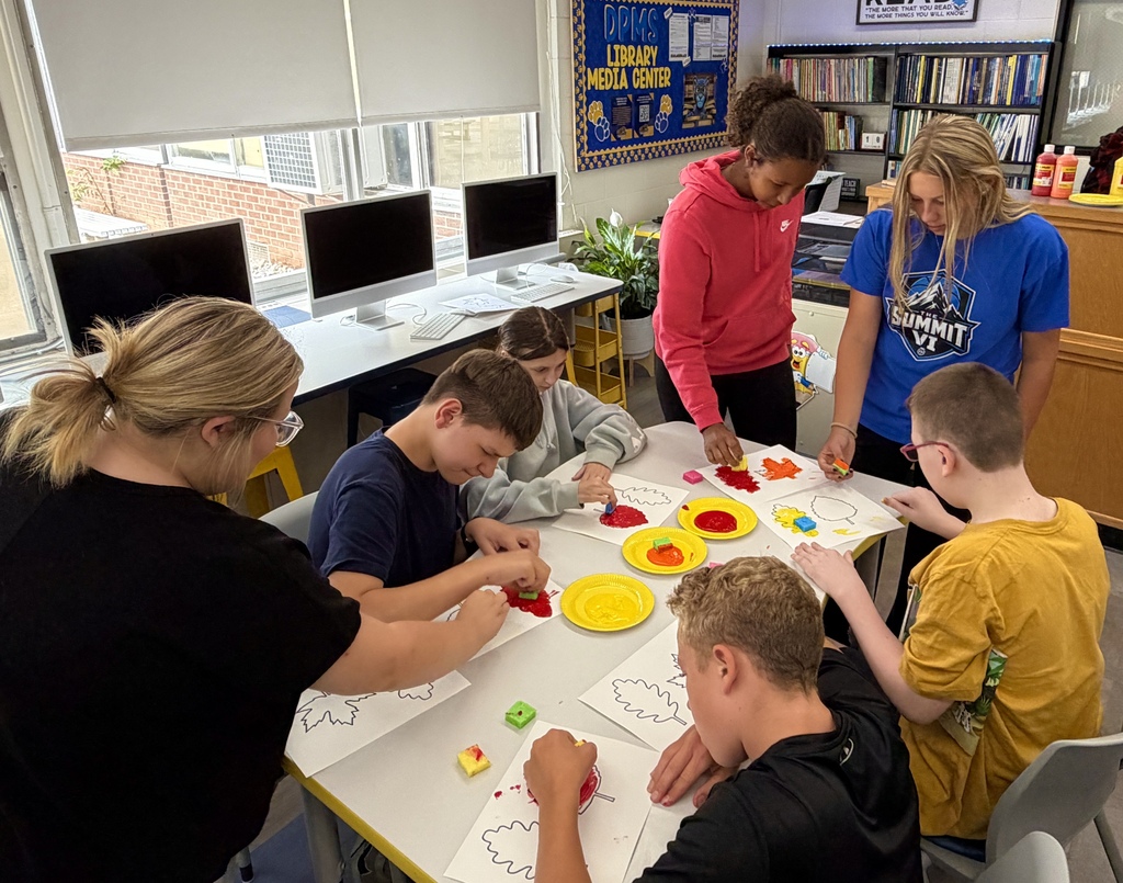 Students are sitting at a classroom table, with some students standing up at the end of the table. They are doing a leaf painting project. 