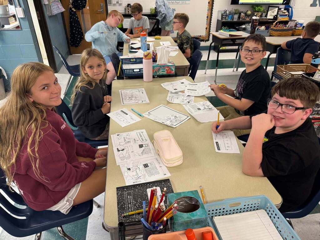 A group of students sitting at a classroom table and looking at the camera. The table has worksheets and other school supplies on it.