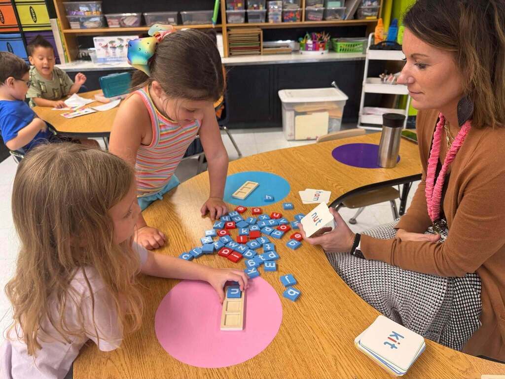 One student is placing a block with the letter n on it inside a tray. Another student is looking through a pile of red and blue letter blocks. The teacher is sitting behind the table and watching the students work.