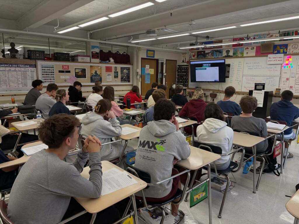 Students sitting in their desks watching a presentation on the TV that is in the front of the classroom.