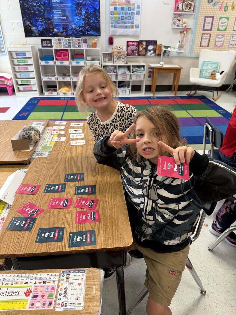 Two students at their desks working through an activity that involves red and black cards. One student is giving the peace sign to the camera and the other is smiling at the student giving the peace sign.