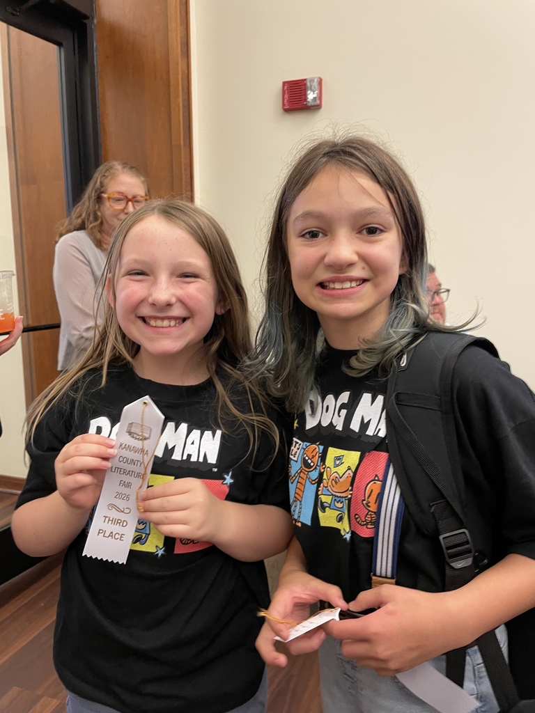 Two smiling girls in matching black graphic T-shirts pose indoors, holding paper tickets; a woman and doorway are visible behind them, creating a cheerful event atmosphere.