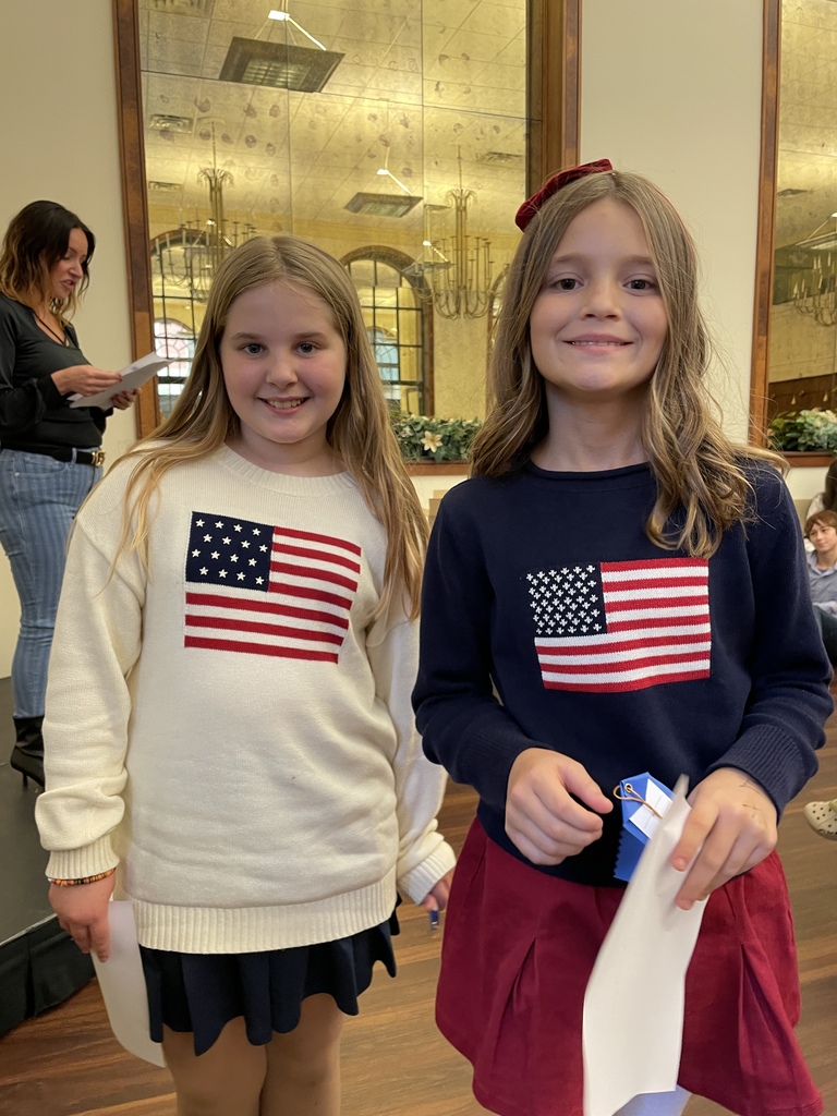 Two smiling girls stand side by side in a bright, ornate room, wearing American flag sweaters; one holds a white garment, creating a cheerful patriotic scene.