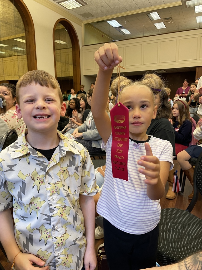 Two children at a school event: a smiling boy stands beside a girl holding up a red award ribbon, with a seated audience in the hall behind them.