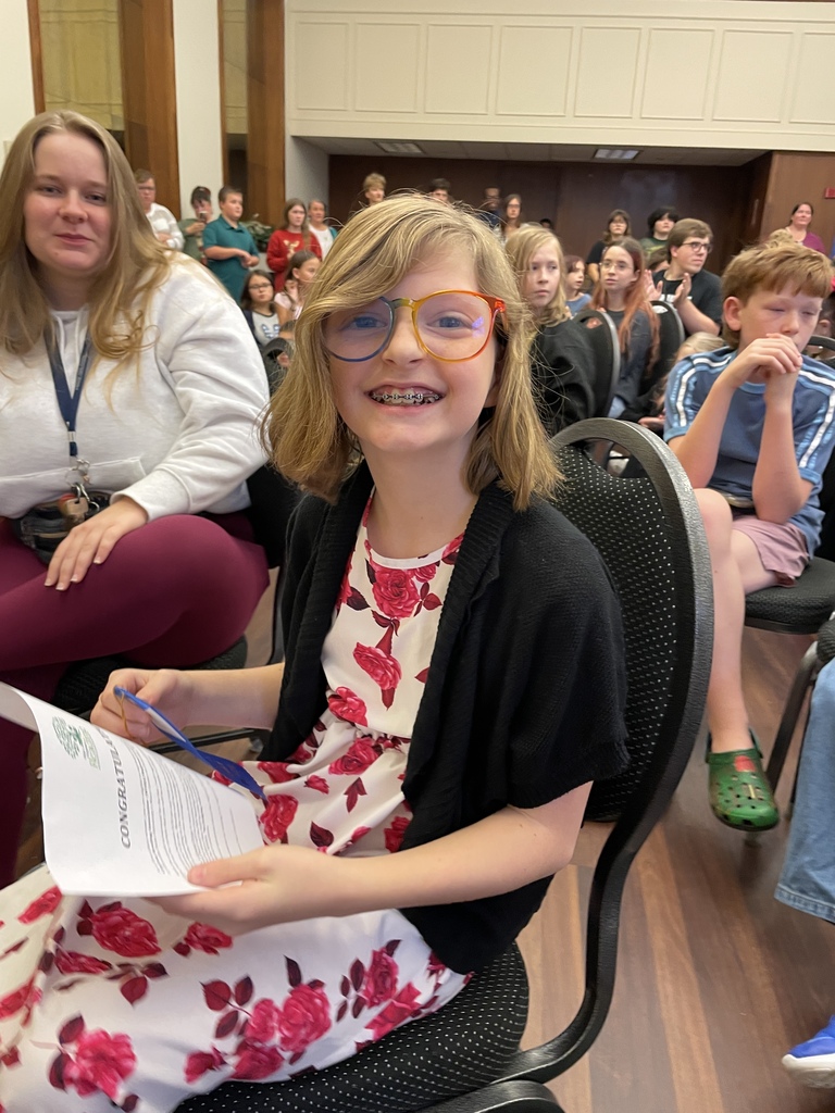 A smiling girl with orange glasses and braces sits in a classroom audience, holding a printed worksheet and pen; students behind her create a lively, welcoming mood.