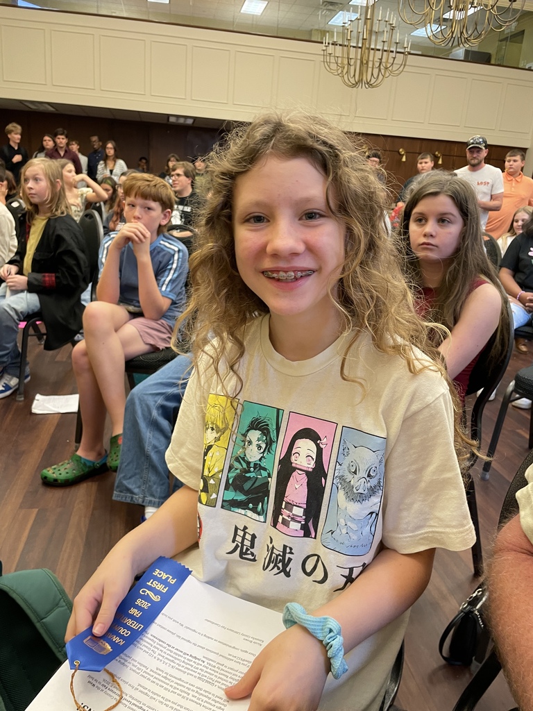 A smiling girl with curly hair and braces holds a blue book in a crowded school auditorium, with rows of seated students and a chandelier behind her.