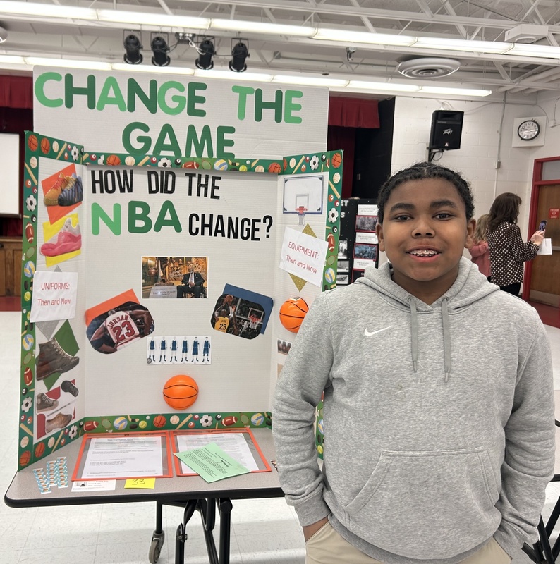 student standing in front of his social studies fair poster. His board is titled "Change the Game" and he asks the question of how did the NBA change through the years.