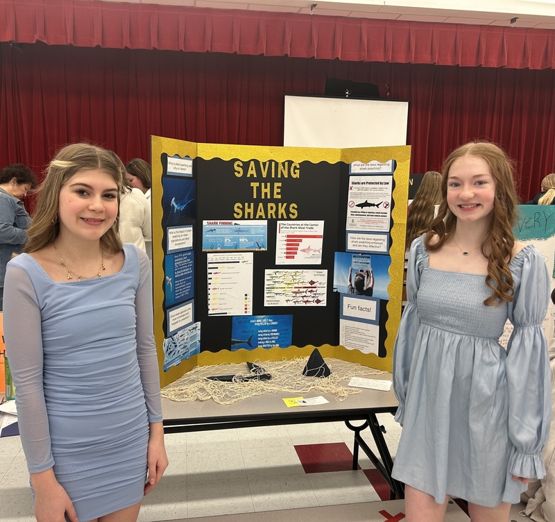 Two students standing in front of their social studies fair project. Their project is titled "Saving the Sharks"