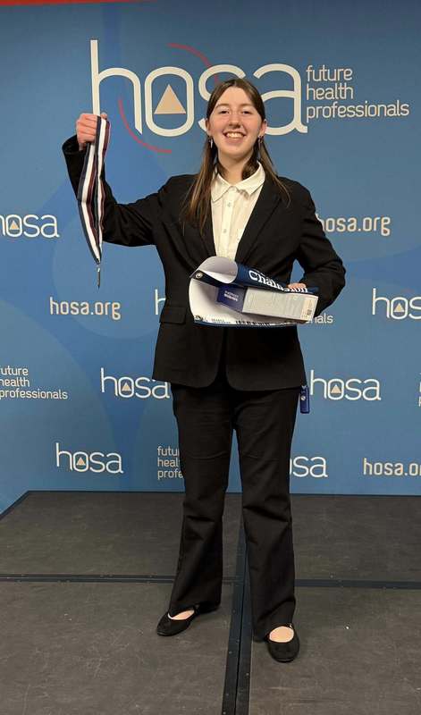 A HOSA student from Ben Franklin is posing for a picture on stage. She is holding up a medal in one hand and is holding onto a championship banner in another hand.