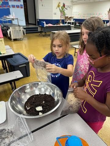 Three students pouring dry baking ingredients into a metal mixing bowl in the school cafeteria. 