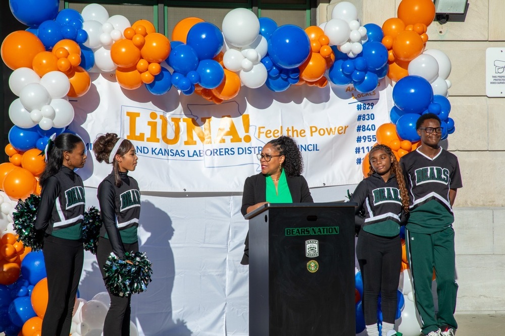 a woman in a green top and black blazer stands behind a podium and is flanked by student cheerleaders. a balloon arch and sign reading "LiUNA" are in the background