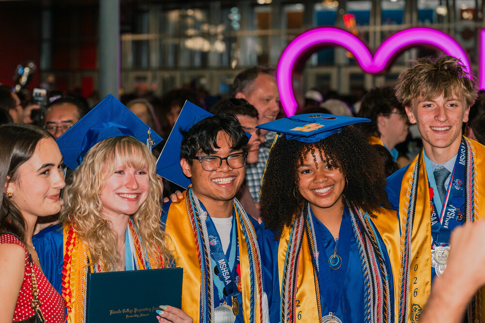 five teens, four of whom wear blue graduation robes and gold stoles with multicolored cords, stand in a line smiling at a camera that is just off screen to the right of the photo