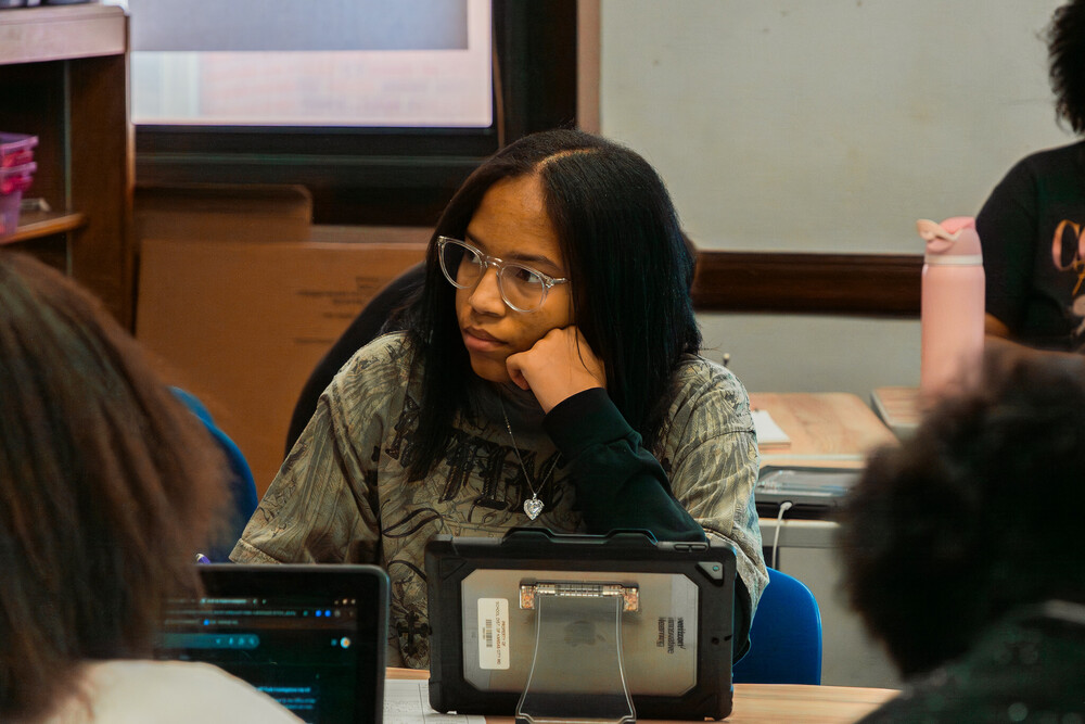 A female student sits at a desk with her chin resting on her fist as she listens to her teacher with an ipad in front of her