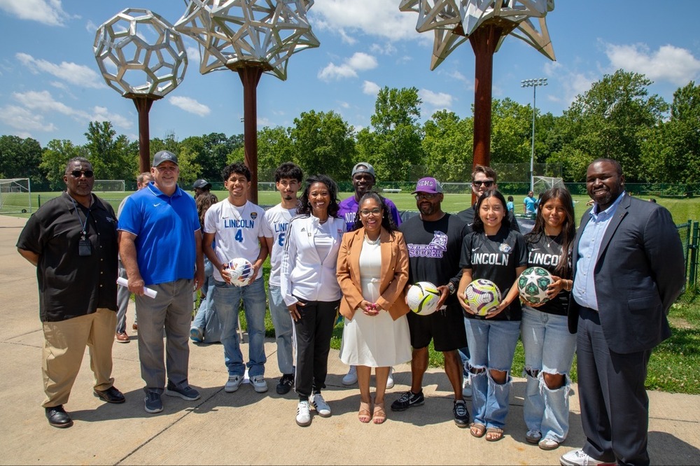 A group of high school student athletes, coaches, and KCPS administrators, including Dr. Jennifer Collier, stand smiling in front of a soccer field