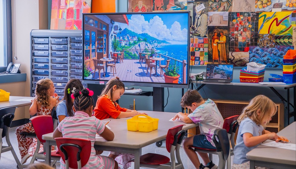 young elementary students sit at classroom tables filling out sheets of paper with pencils
