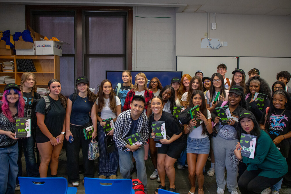 A large group of high-school aged students stand in a group holding "wicked" books and smiling at the camera