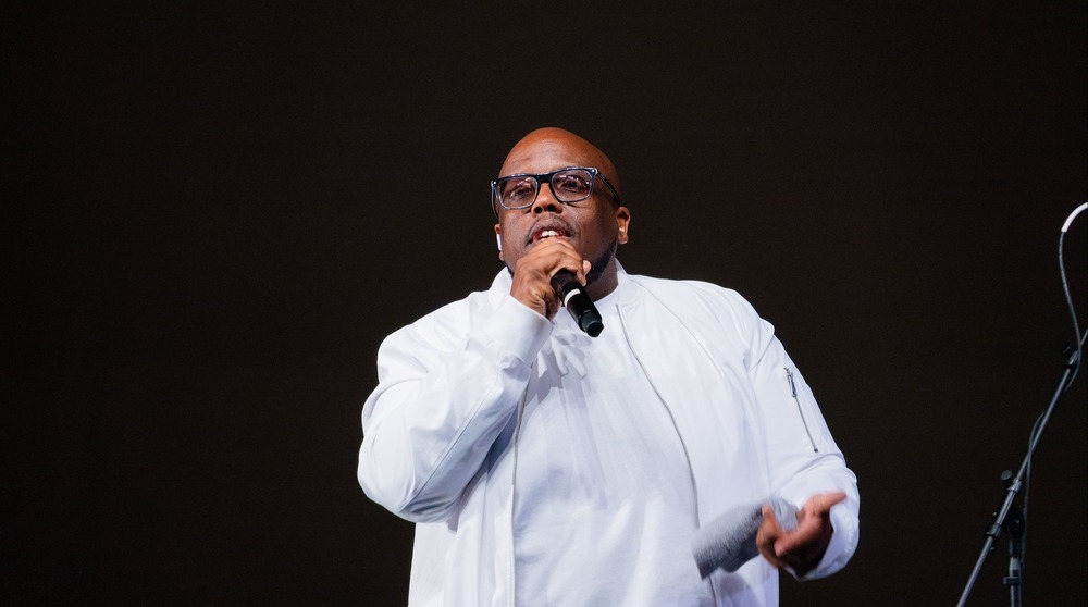 man in a white t-shirt and jacket stands on stage holding a microphone as he addresses a crowd
