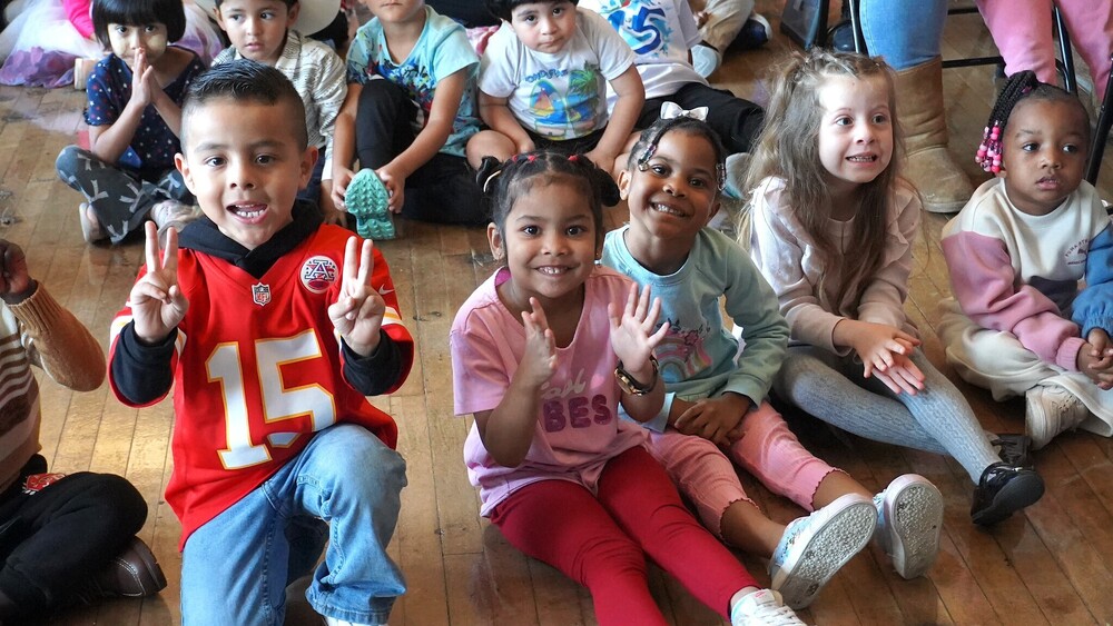 A group of 3-5 year old children sit on a gym floor smiling at the camera