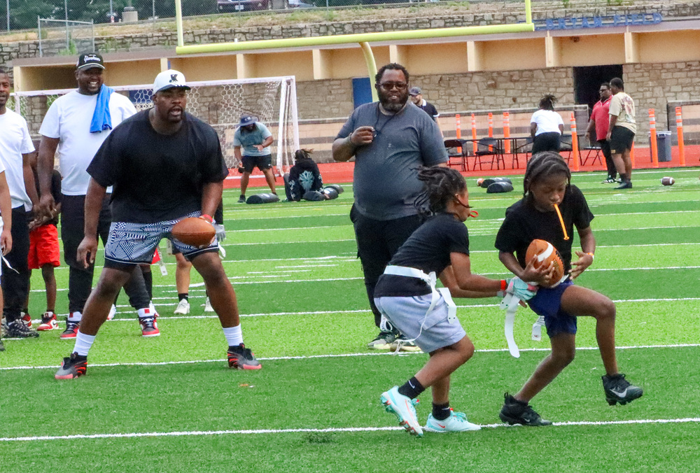 Two young children play flag football in the foreground of the photo while several men, including Chiefs player Chris Jones, stands in the background watching and coaching them.