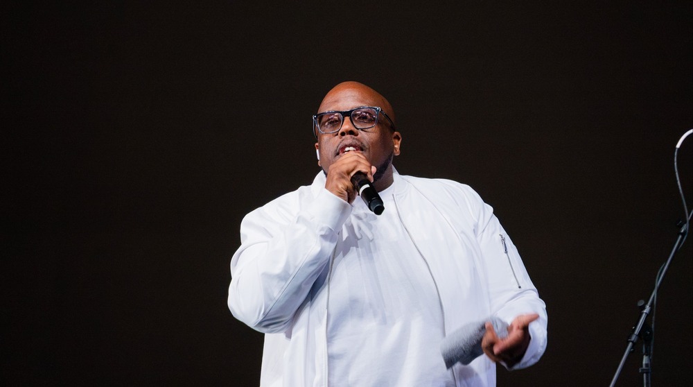 man in a white t-shirt and jacket stands on stage holding a microphone as he addresses a crowd