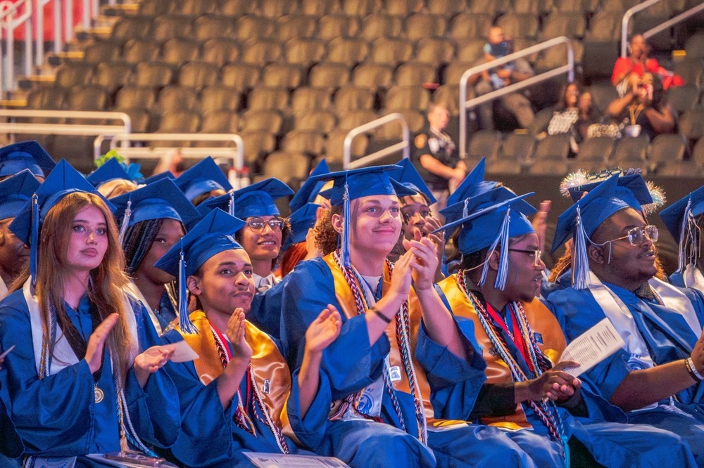 A group of students in blue graduation caps and gowns are seated and looking forward as they applaud.