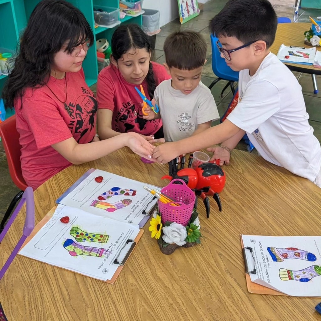 Two older students interact with two young students as they sit at a classroom table with toys on it.
