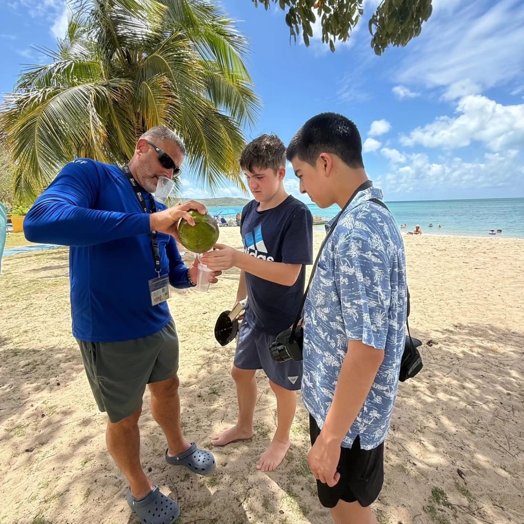 Two students watch as a teacher pours fresh coconut water from a coconut into a cup. They are standing on a beach with a palm tree in the background.