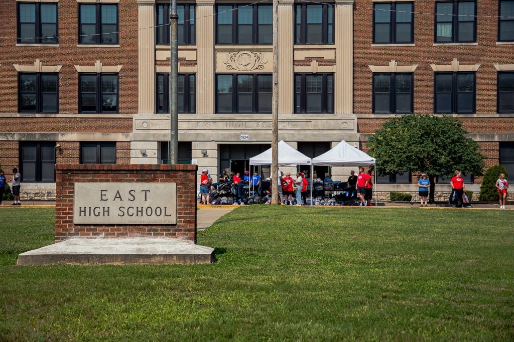 A large brick school building stands in the background, with a sign reading "East High School" in front.