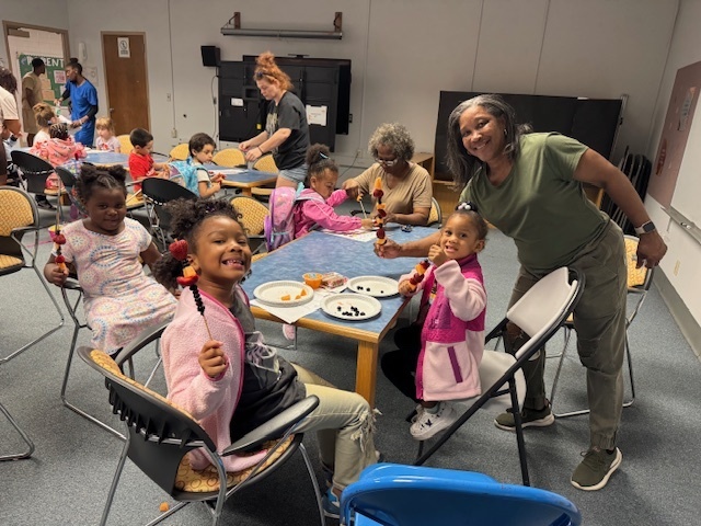 A group of preschool children with a teacher eating fruit kabobs
