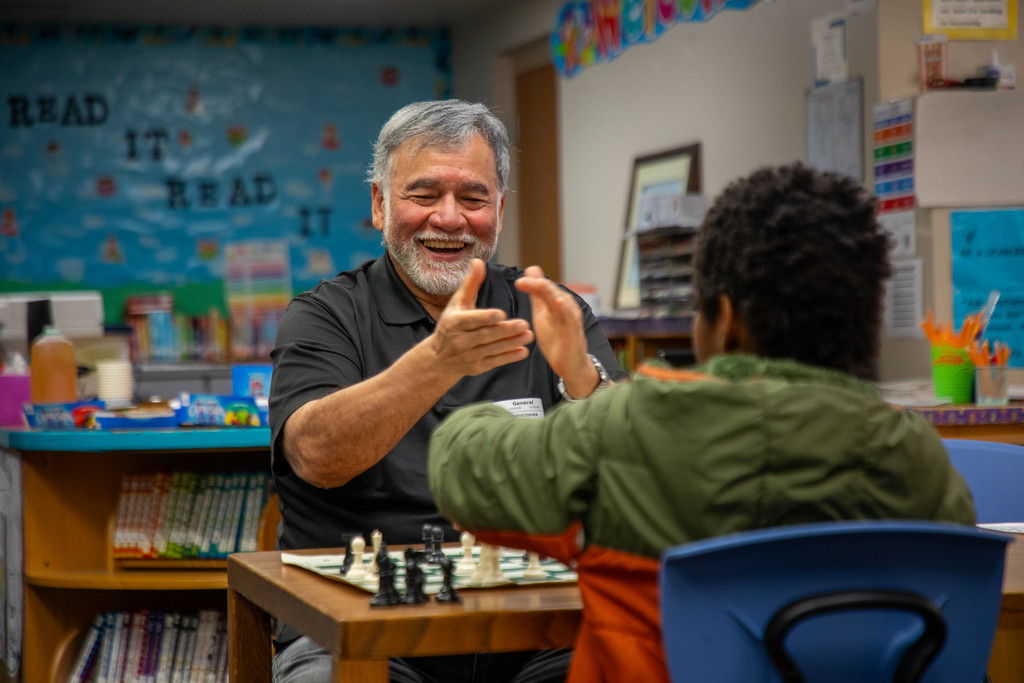 Man claps as smiles as he sits across from a young student at a chess board