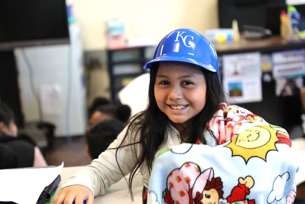 A young student smiles at the camera wearing a Kansas City Royals blue plastic baseball helmet