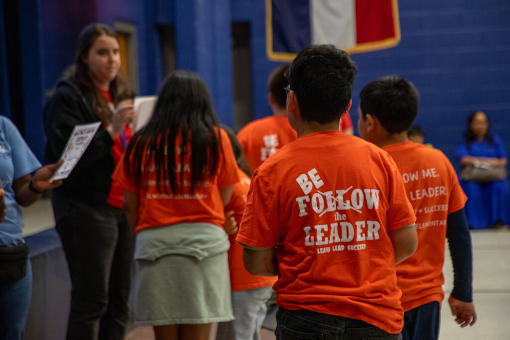 Students wearing matching orange shirts that say "be the leader" on the back are lined up to receive academic achievement certificates