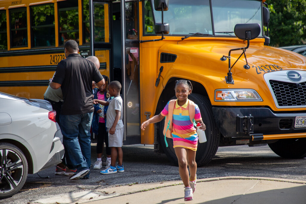 A student in a rainbow-colored dress runs from a school bus toward the school with a smile on her face